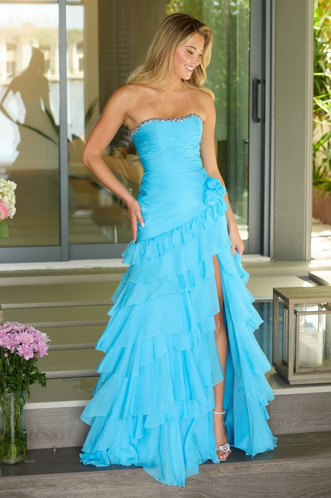 A woman in the strapless Ava Presley 42242 ruffled prom gown in sky blue stands on steps by a glass door, holding the side of her evening dress and smiling.